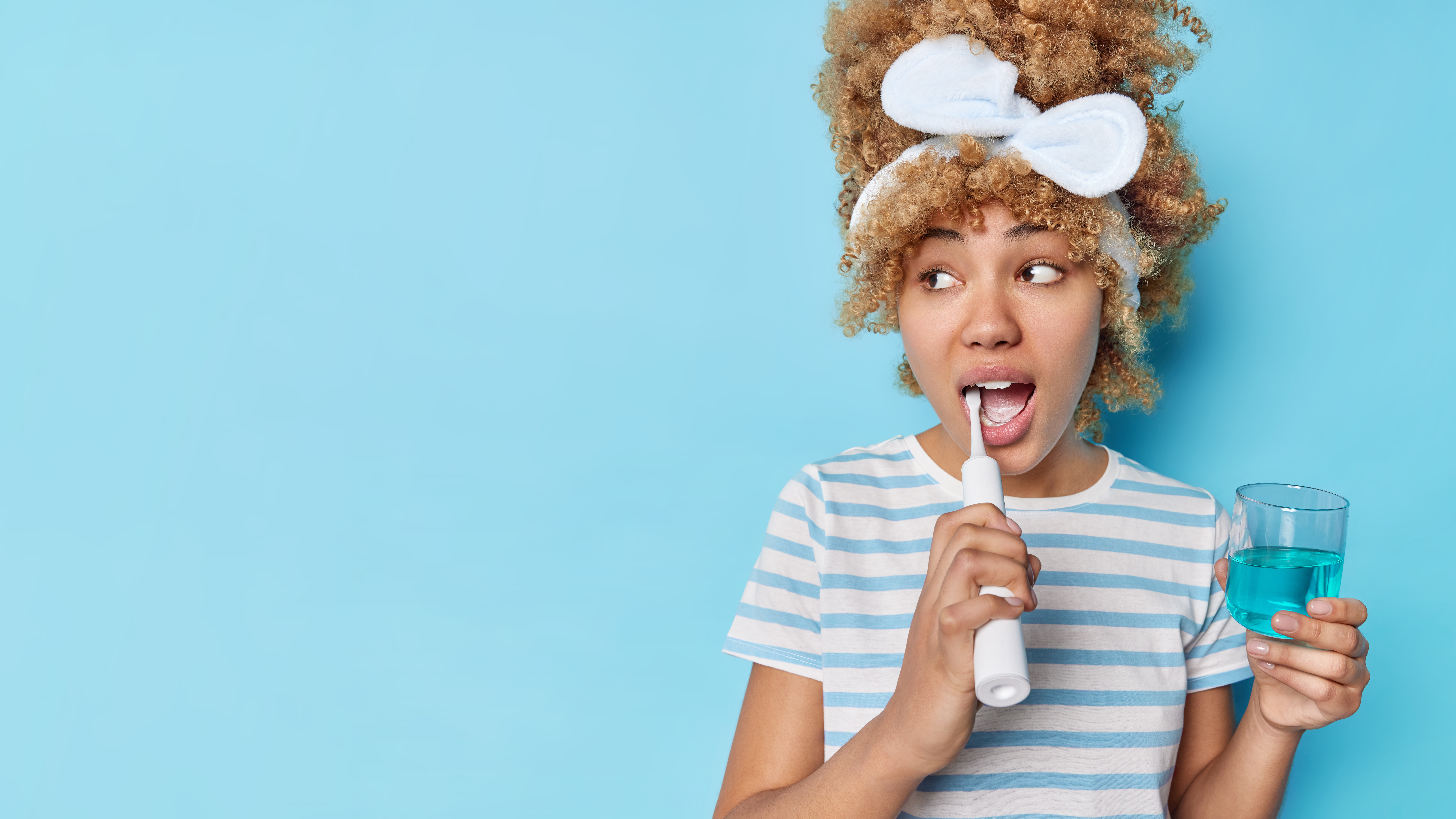 Photo Blonde Curly Woman Brushes Teeth With Electric Brush Holds Glass Mouthwash Fresh Breath Isolated Blue Background Blank Space Your Promotional Content Dental Care Concept